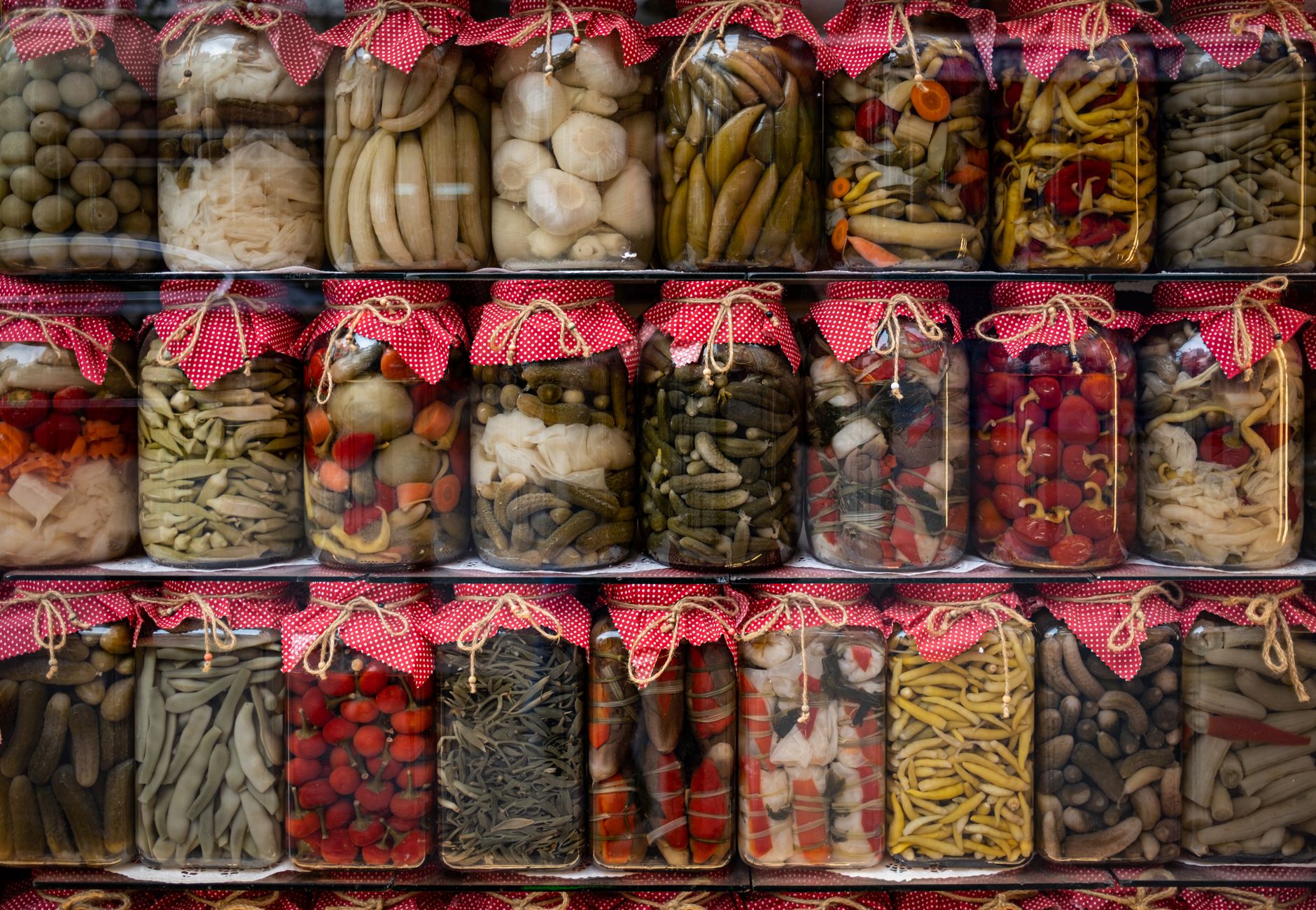 Vibrant assortment of pickled vegetables in jars with red fabric covers, displayed on shelves.