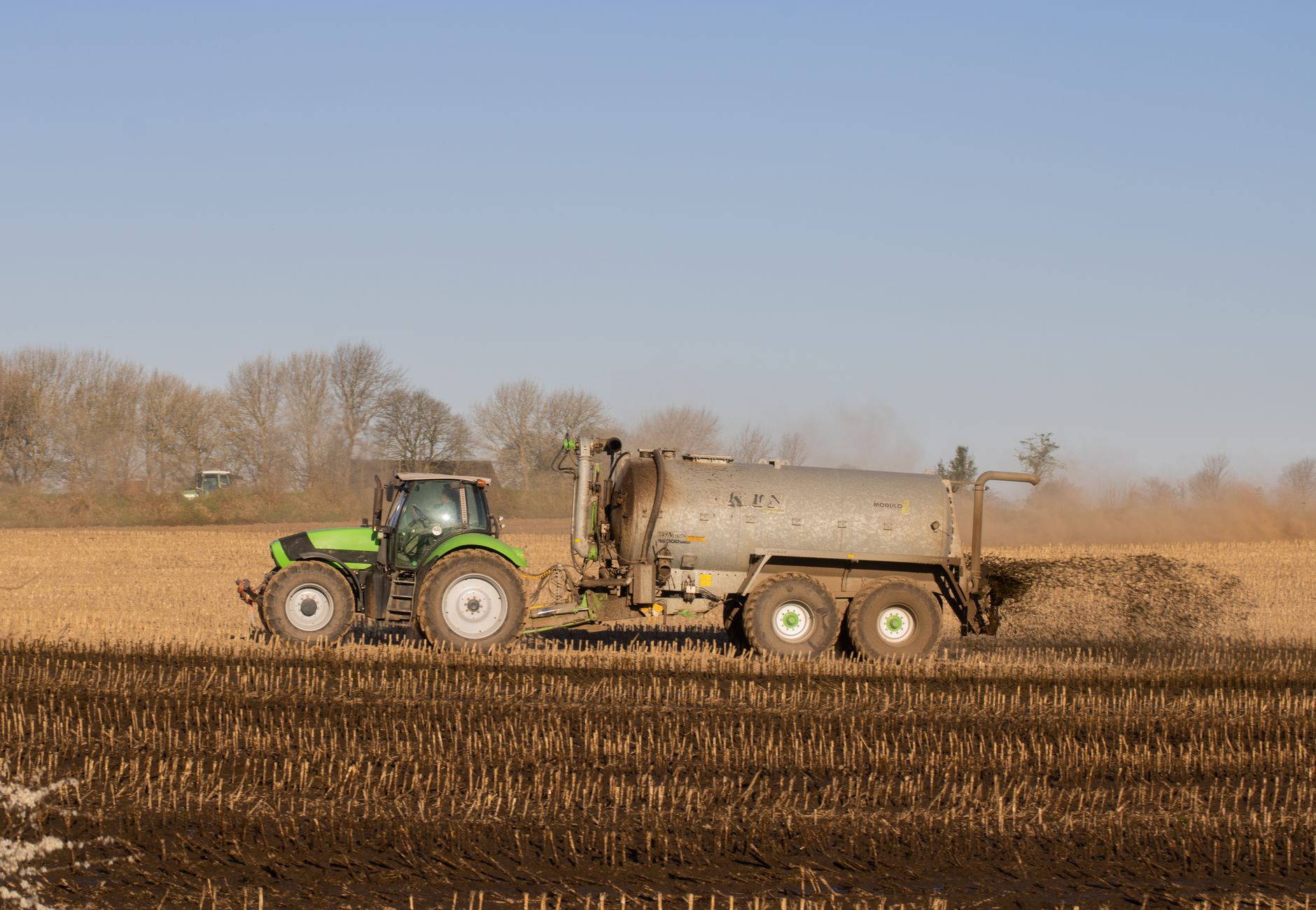 A tractor with a tanker spreading fertilizer over a barren farmland under clear skies.